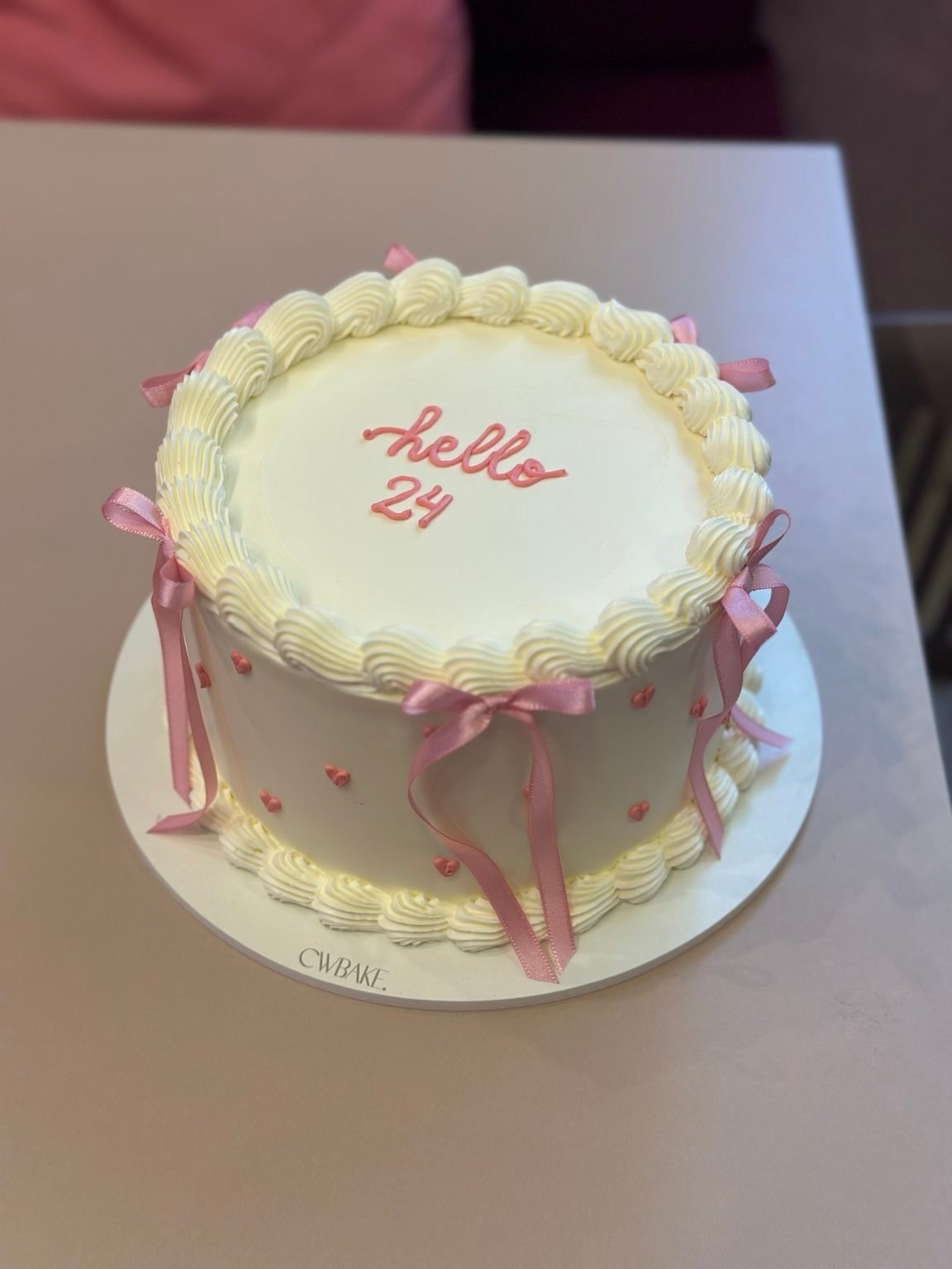 Bolo vintage branco com laços cor-de-rosa e a frase “hello 24”