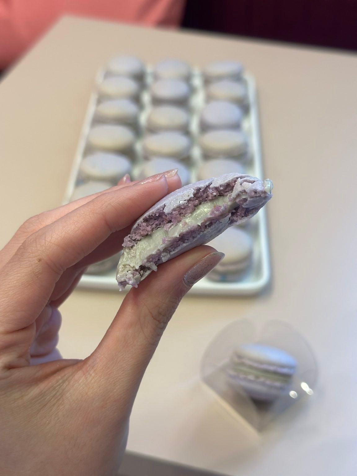 Mão segurando um macaron recheado, com bandeja de macarons ao fundo